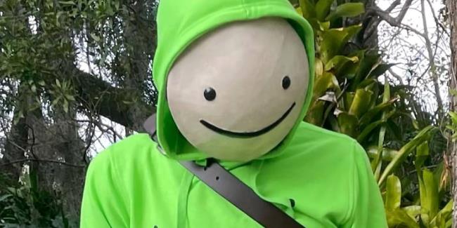 Dream YouTuber standing in front of trees in a lime green hoodie with his ico<em></em>nic white circular mask with a smiley face painted on top