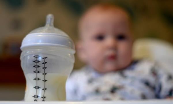 A baby in a high chair looking towards her bottle of milk in the foreground