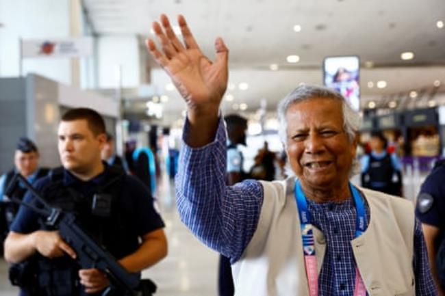 An older Asian man waves in an airport as armed men stand behind him