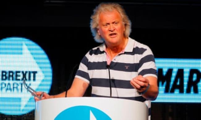 Tim Martin standing at a podium making a speech surrounded by the pale blue graphics and livery of the Brexit party