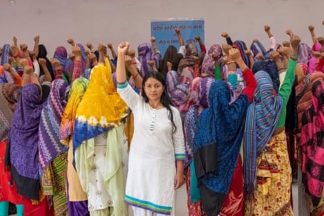 Devi Khadka faces the camera at the centre of a crowd of women dressed in saris with their backs to the camera. All have one hand raised in a fist