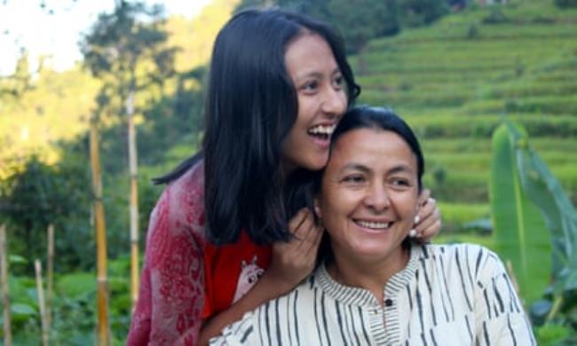 Two women smile looking at something in the distance. The younger woman is standing behind the older woman and lightly cradles her head