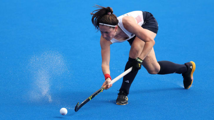 brown-haired woman a<em></em>bout to hit a field hockey ball with her stick; water splashes from the field