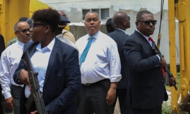 An older African Caribbean man in a tie stands surrounded by guards in suits holding assault rifles  