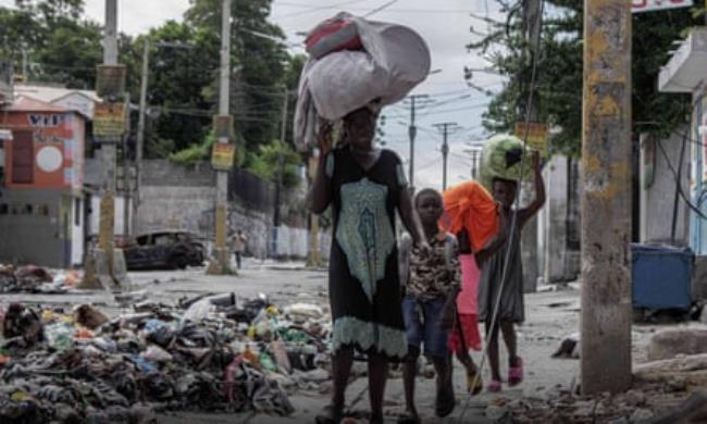 An African-Caribbean woman and three children carry bundles of belo<em></em>ngings as they walk past piles of rubbish in a street with a burned-out car int the background