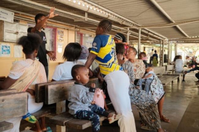 A small group of women and children sit on benches under a tin roof while a medical worker talks.