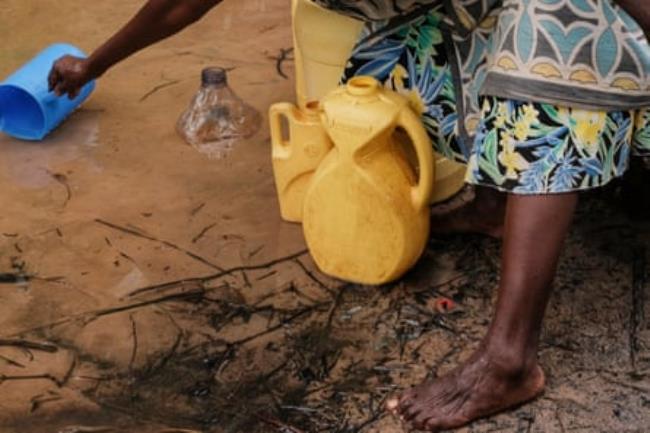 A woman reaches into cloudy water with a blue jug in order to fill two yellow water co<em></em>ntainers at her feet.