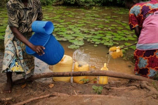 Two women stand on a muddy bank and fill brightly coloured buckets and co<em></em>ntainers 