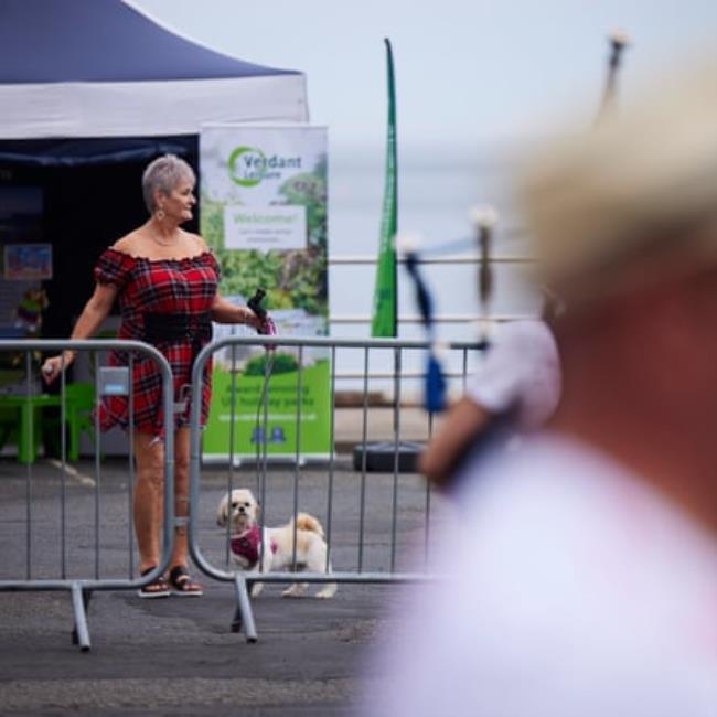 An older woman in a short tartan dress watches a pipe band pass with her small white dog, which also seems to wear tartan 