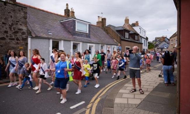 A procession of adults and children, led by women in sashes, walk down a road of small houses