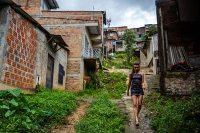 A trans woman walks down a narrow path between houses