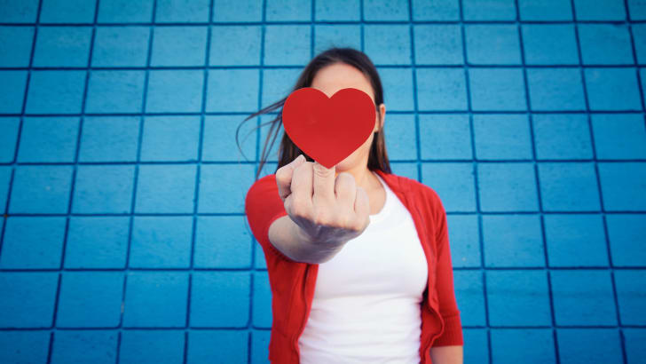 woman stands in front of a blue wall and flips off the camera while co<em></em>ncealing it with a paper heart