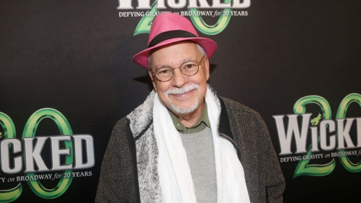 bespectacled man with white, close-cropped facial hair; wearing a pink hat and green button-down beneath neutral outerwear