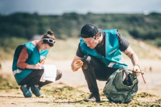 Two volunteers wearing blue tabards crouch on a beach. One has a sheaf of paper and is writing, the other has a bag and is examining something he has picked up.