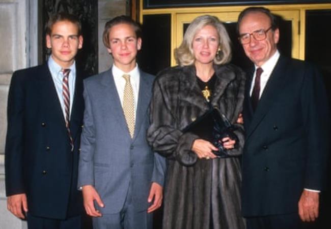 Color photo of four white people standing in a row, the men wearing suits and the woman wearing a dark gray fur coat. Two men are young, one is older.