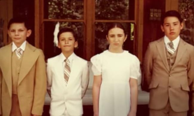 A deliberately grainy 80s-era image of four white kids, in beige and white, the boys in suits and ties and the girl in a dress, standing outside in front of French doors.