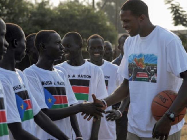 A very tall young African man holding a basketball shakes hands with a row of teenage boys wearing T-shirts with the South Sudan flag 