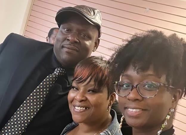 A man in a suit wearing a leather cap poses for a selfie with two smiling women. 