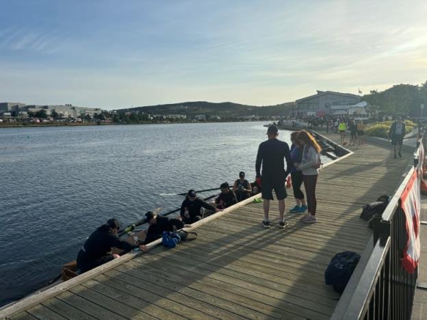 Shoreline of lake with people standing on board as people sit in a boat