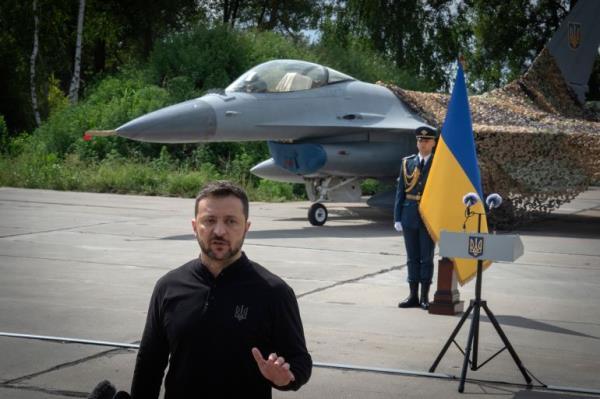 Volodymyr Zelenskyy pictured speaking at an airba<em></em>se at an undisclosed location. There is a Ukraine flag behind him and an F-16 fighter jet