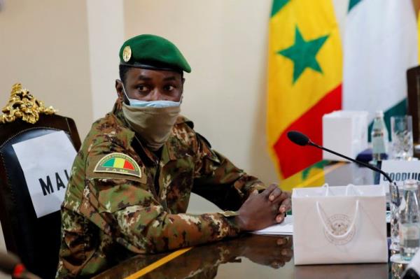 Goita sits at table, wearing military uniform and a green cap.