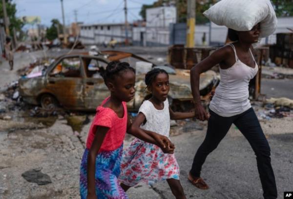 FILE - Girls holding hands are led past a burnt car blocking the street as they evacuate the Delmas 22 neighborhood to escape gang violence in Port-au-Prince, Haiti, May 2, 2024.