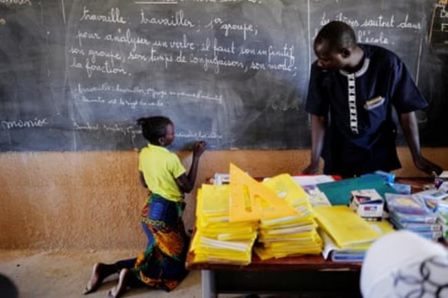 A girl writes on a blackboard while a teacher watches