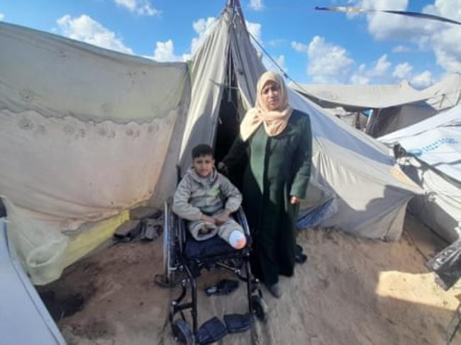 A woman stands next to a boy in a wheelchair outside a tent.