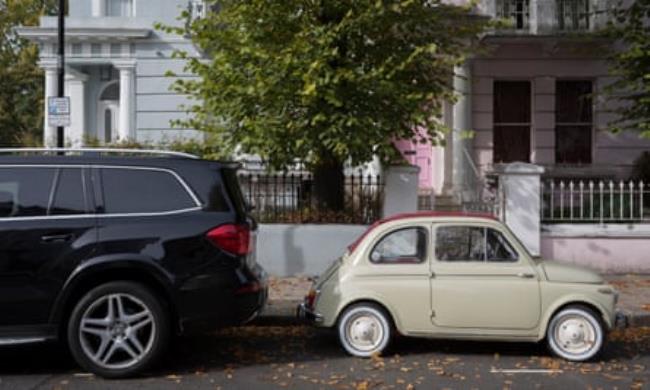 A large 4x4 SUV car parked behind a much smaller Fiat 500 among autumn leaves on Elgin Crescent in Notting Hill